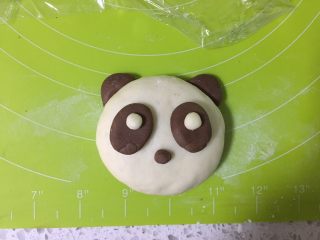 11、呆萌熊猫豆沙包,另取两个可可小面团，搓成椭圆形，按扁，沾一点水沾在熊猫的脸上，白面团粘在眼袋上，再取一点点可可面团作为鼻子