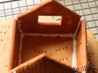 16、姜饼屋,将烤好的姜饼放在底座上。首先拼装底部，将装有蛋白糖霜的裱花袋用剪刀剪一个小口，用糖霜把前、后及侧墙这四块饼干粘起来