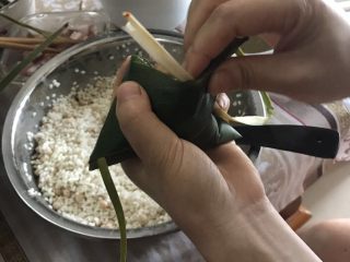 8、肉粽子,把粽子叶往手肘的这边收，收的时候先收左边，保证左边糯米全部被粽叶包住，之后收右边；之后把多出来的粽子叶往右边放回去，放之前把棕叶的根部夹在粽子叶中间（这样捆粽子的棕叶两头都跟粽子连在一起啦）