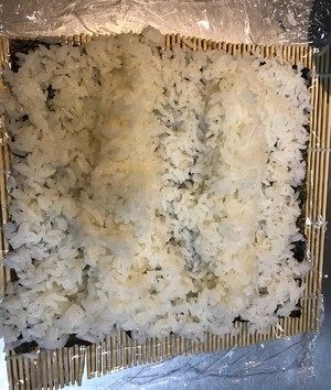 1、花朵寿司,寿司帘平铺保鲜膜，保鲜膜再铺海苔，海苔上再铺米饭，用手按压平整。在中间部分再用米饭筑两道墙。