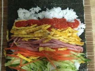 3、紫菜包饭,取紫菜铺竹帘上，放上调上寿司醋，盐，糖的米饭，加上之前切好的配料，如图