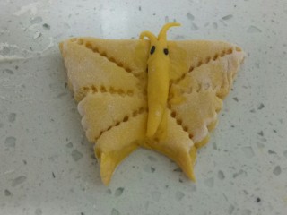 9、南瓜花样小馒头,取一个小剂子，先揉光滑，速度要快否则会发酵过度。用擀面杖把面团擀成椭圆形的牛舌状，从中间折起来，中间稍粘一点水，可以稍微整一下形，用寿司卷帘从中间压下去，再从上面和下面压，用锋利的刀把下面切下来，搓成蝴蝶的身体，用清水粘在翅膀上，最上面切个嘴，用黑芝麻粘上眼睛点缀。用刮刀在身体两侧按出一些花纹，用纯奶吸管在身体上按上圆圈。