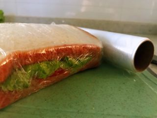 6、鸡胸肉三明治,用保鲜膜紧紧的裹起来,全部包裹完之后,拿刀从中间切开,此时,案板和刀特别重要了,为了避免细菌的交叉感染,生熟案板板和刀一定是分开的。