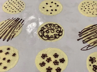 4、烟卷饼乾,将可可面糊袋前端剪一小洞(小一点画出来线比较细)，画上喜欢的图案。完成后放入烤箱以170度烤7-9分，观察边缘呈金黄色泽即可。烤箱关掉，一次取一片出炉，最好能在烤箱旁边操作，出炉立刻翻面，然后用筷子捲起定型，捲到尾端时稍微压一下固定，再继续取出饼乾片捲下一个。