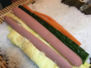 6、寿司卷,将适量米饭放在事先铺好的寿司帘的海苔上面，带上一次性手套按压结实，最上面留出一些空间。