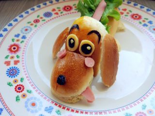 18、小热狗造型面包,用海苔和奶酪片刻出眼睛，眉毛，蘸少许沙拉酱粘在相应的位置。一粒黑提子干做鼻头，再在背部的香肠上挤上沙拉酱就可以了。