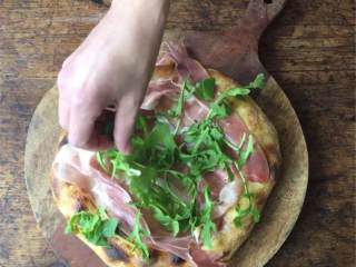 7、芝麻菜熏火腿披萨,淋上特级初榨橄榄油 ，撒上海盐 。把披萨，烘焙纸等一起转移到预热好的铁盘上，烤5分钟，然后从烤箱中取出，撒上意大利熏火腿 和芝麻菜 。