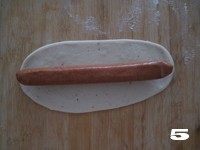 5、【花边培根披萨】换着花样吃披萨,取另外1/3面团揉匀擀成牛舌状饼皮，将香肠完全包裹在内，用刀切成均匀的片状。